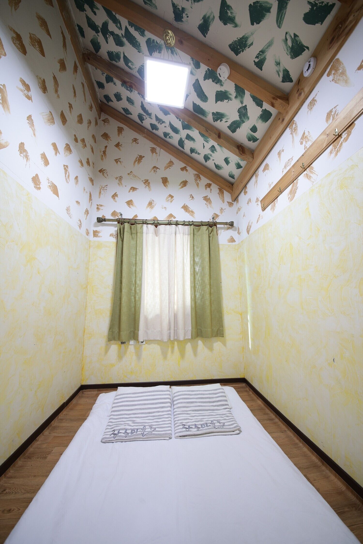 A room with a bed, wooden beams, patterned wallpaper, a window with curtains, and a ceiling light.