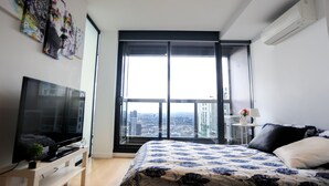 Une chambre moderne dotée d’une grande fenêtre, d’un téléviseur à écran plat, d’un lit avec une literie à motifs et d’une petite table sur laquelle est posé un vase.