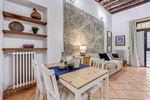 A dining area with a table, chairs, a radiator, shelves with decorative items, a large patterned wall, and a sofa.
