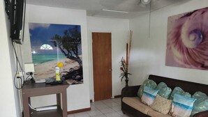 A living room with a beach-themed poster, a television, a sofa with patterned cushions, a decorative plant, and a tiled floor. - Sagusta Gardens NCA 1 (Whitby)
