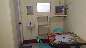 Ein kleines Zimmer mit Tisch, Stuhl, Waschbecken, Fenster und Fernseher.