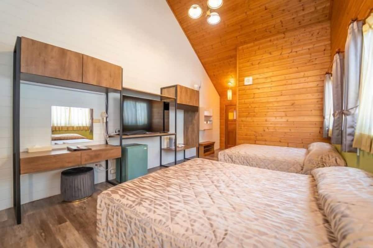 Quarto com teto inclinado, paredes de madeira, cama com roupa de cama estampada, escrivaninha com computador e uma pequena lixeira.