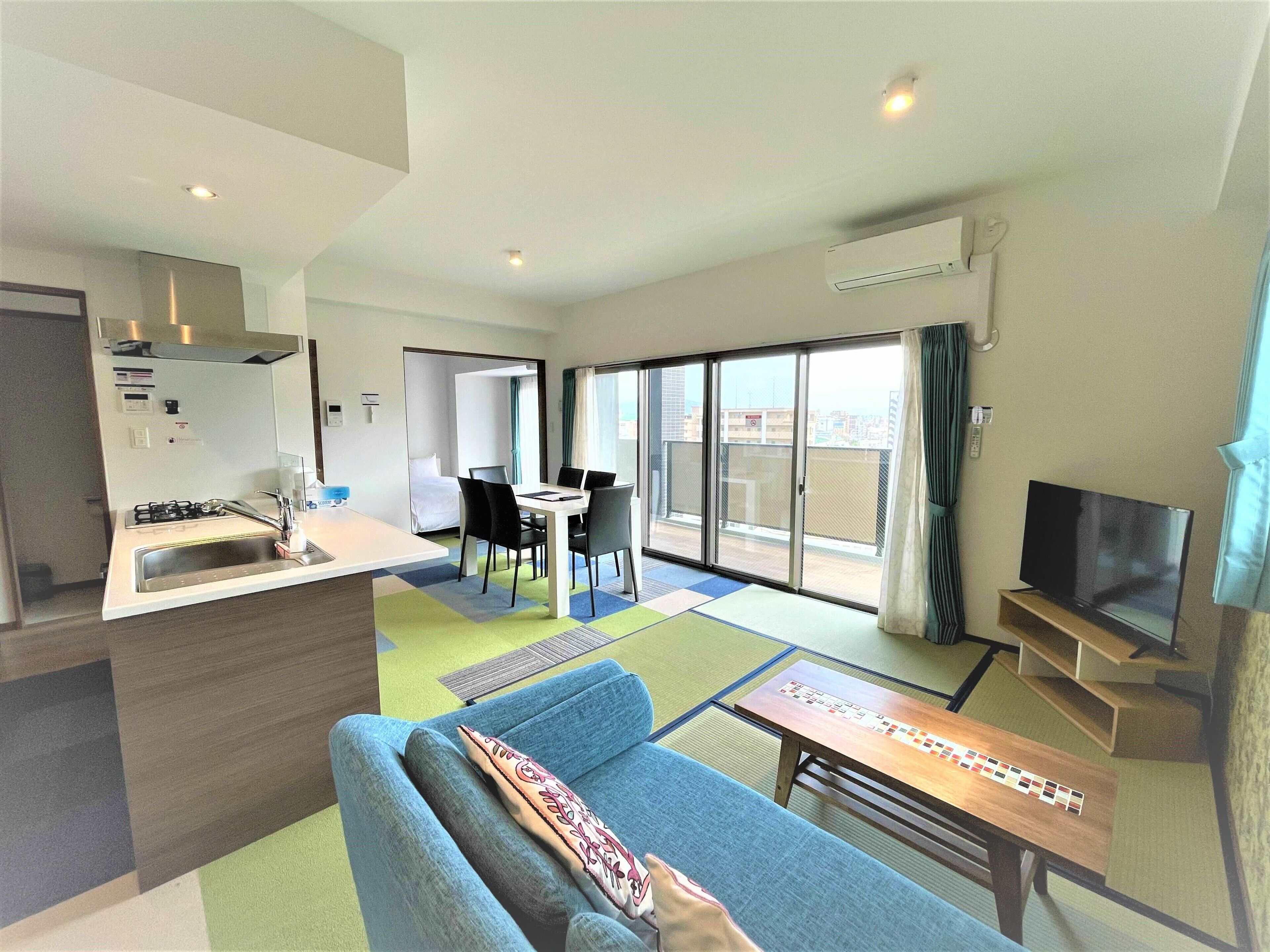 A modern hotel room with a kitchenette, a dining area, a sofa, a TV, and a balcony with a city view.