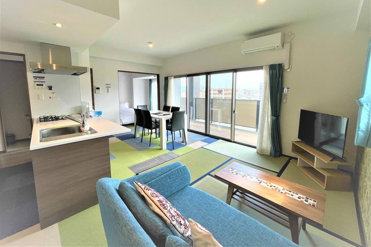 Una habitación de hotel moderna con kitchenette, zona de comedor, sofá, televisor y balcón con vista a la ciudad.