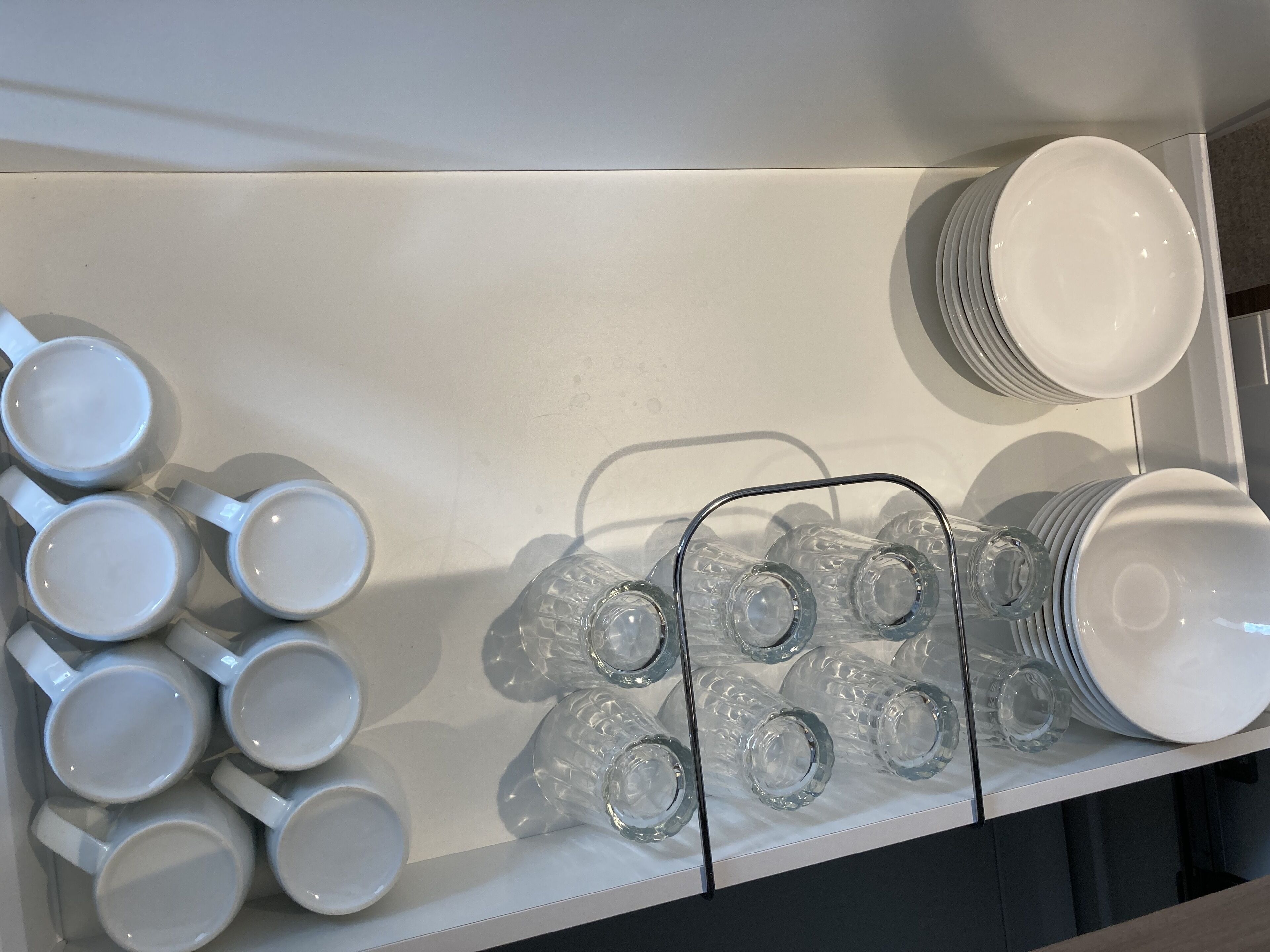 A white shelf with neatly arranged white mugs, clear glass tumblers, and stacked white plates.