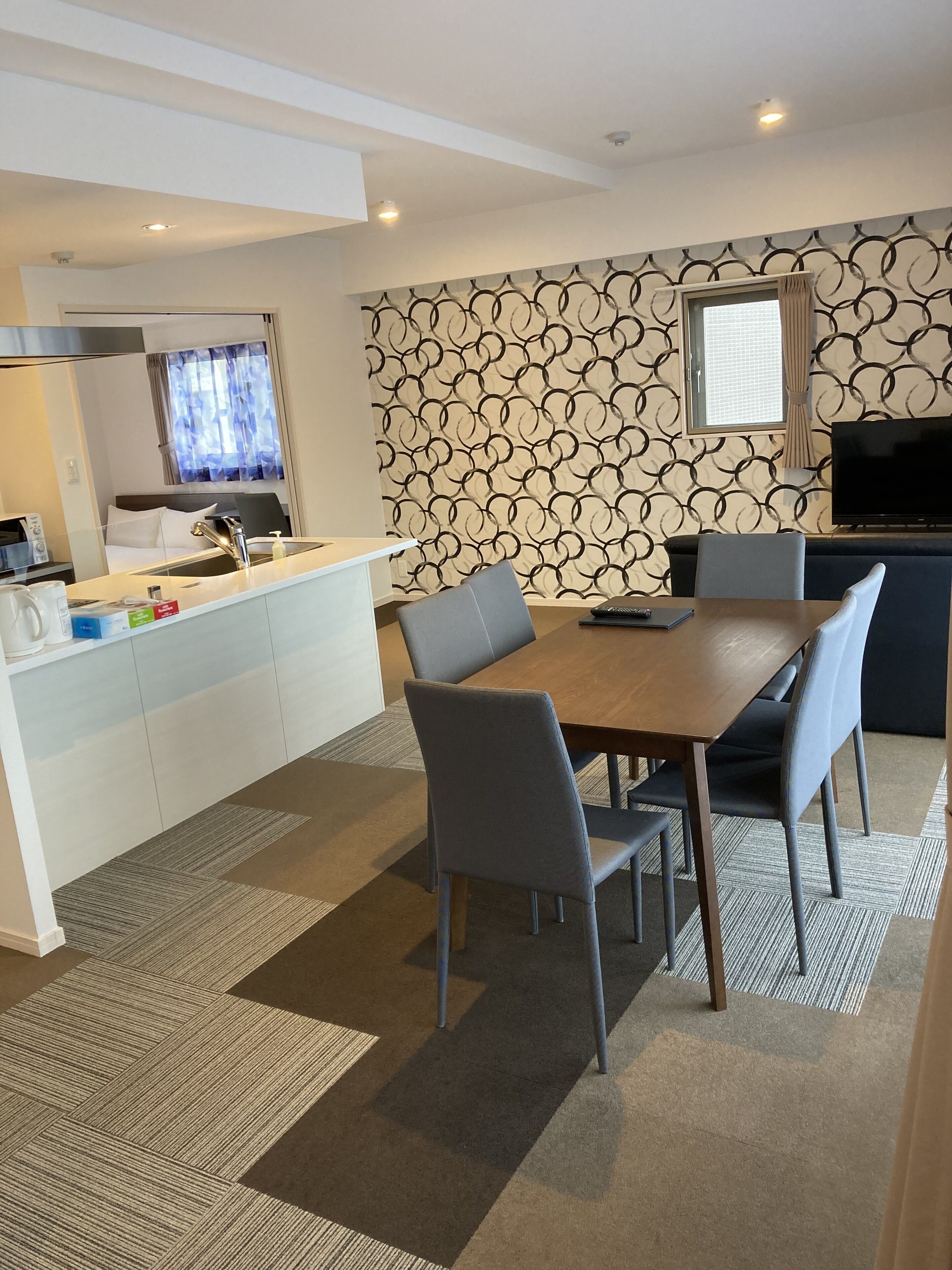 A modern kitchen with a built-in stove, a dining area with a wooden table and chairs, and a patterned wallpaper.