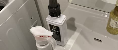 A bathroom sink with a white bottle of hand sanitizer, a small bottle of foam soap, and a mirror above the sink.