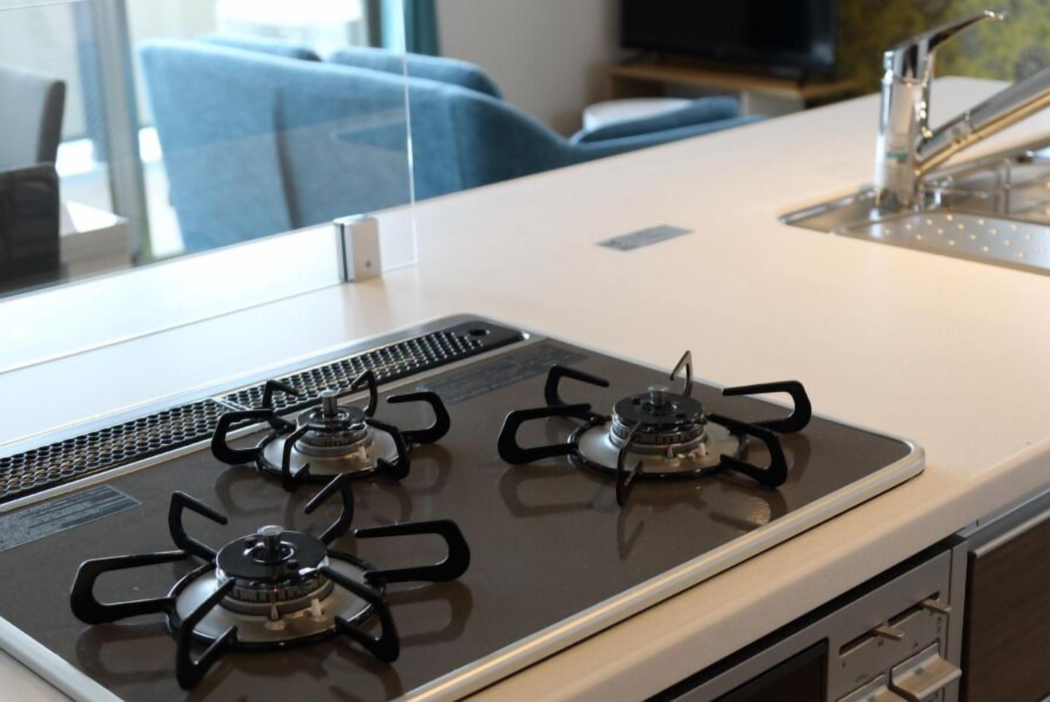 A kitchen with a gas stove, a sink, and a window with a view of a blue chair and a television in the background.