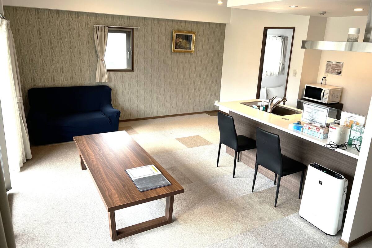 Habitación de hotel compacta con sofá, una pequeña mesa de comedor, kitchenette y un basurero.