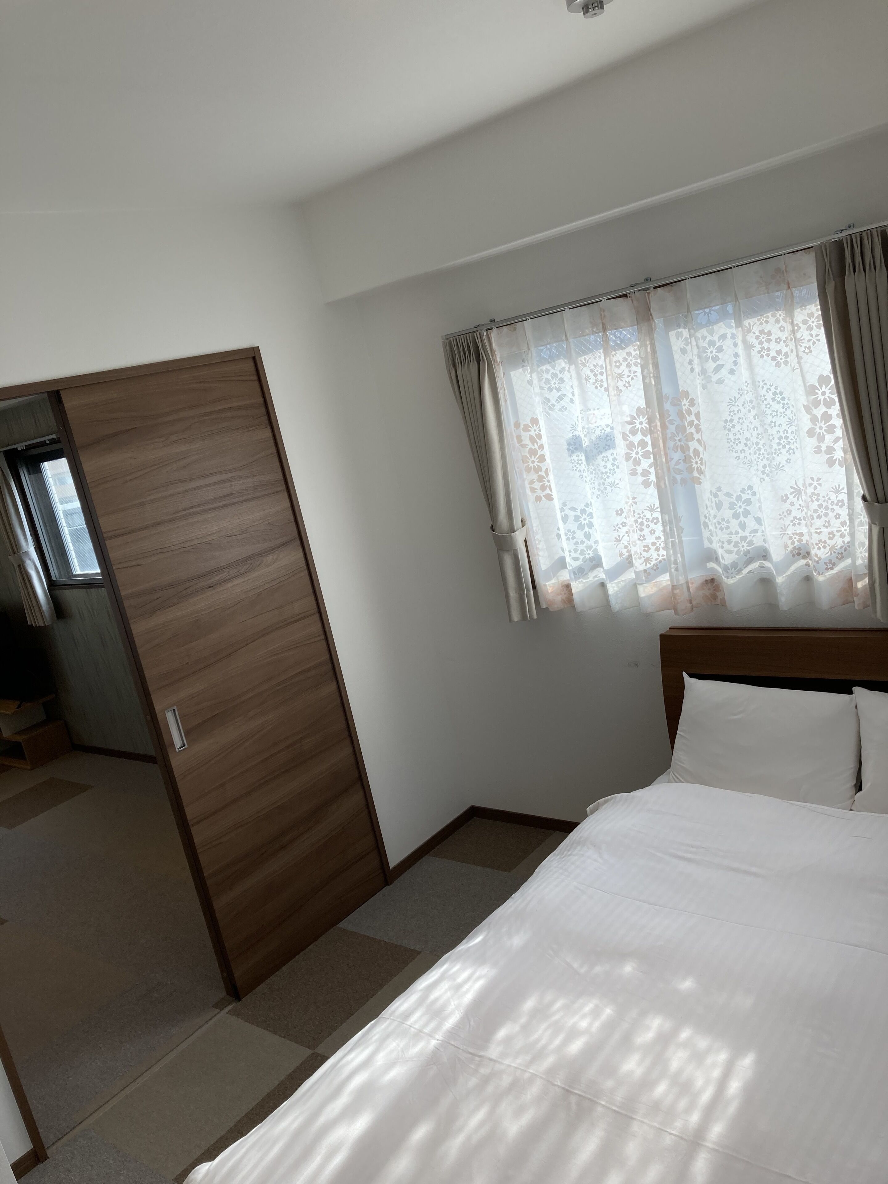 Une chambre d’hôtel avec une armoire en bois, un lit avec du linge de maison blanc et une fenêtre agrémentée de rideaux à motifs floraux.