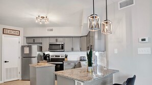 Une cuisine moderne avec des meubles gris, un réfrigérateur en acier inoxydable, un four à micro-ondes et une cuisinière. On y trouve deux lampes suspendues, un petit îlot central avec un évier et une chaise noire.