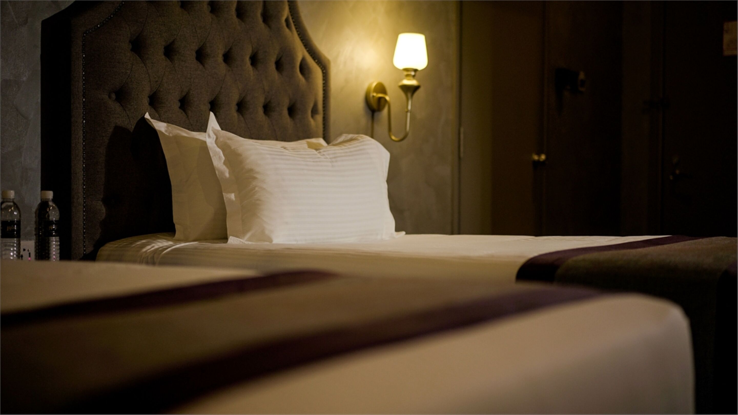 Habitación de hotel con cabecera capitonada, lámpara de noche y camas bien hechas.