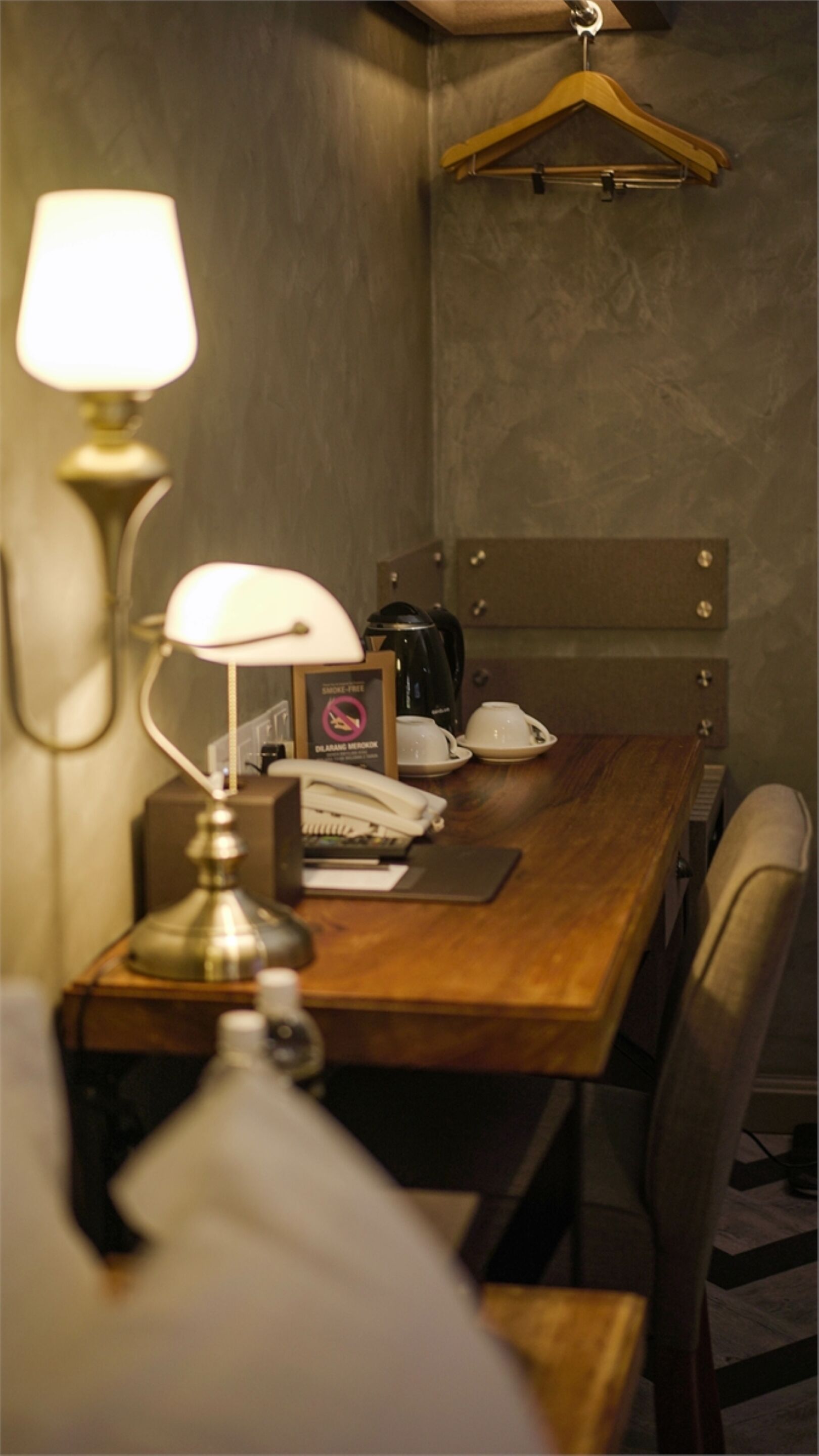 A hotel room with a wooden desk, a lamp, a telephone, a coffee maker, and a hanging coat rack.