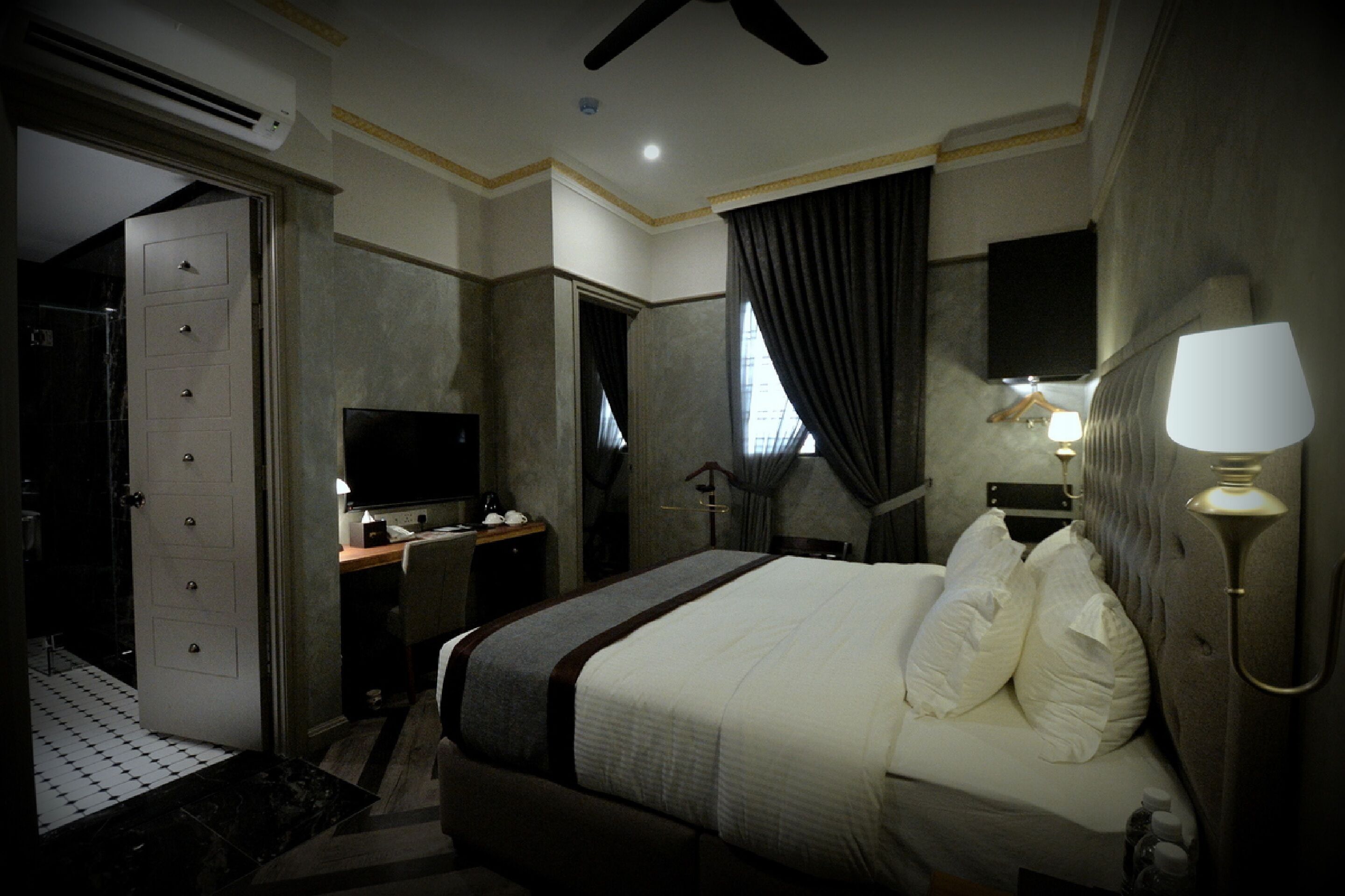 Habitación de hotel con cama, escritorio, televisión, lámpara y baño con ducha e inodoro.