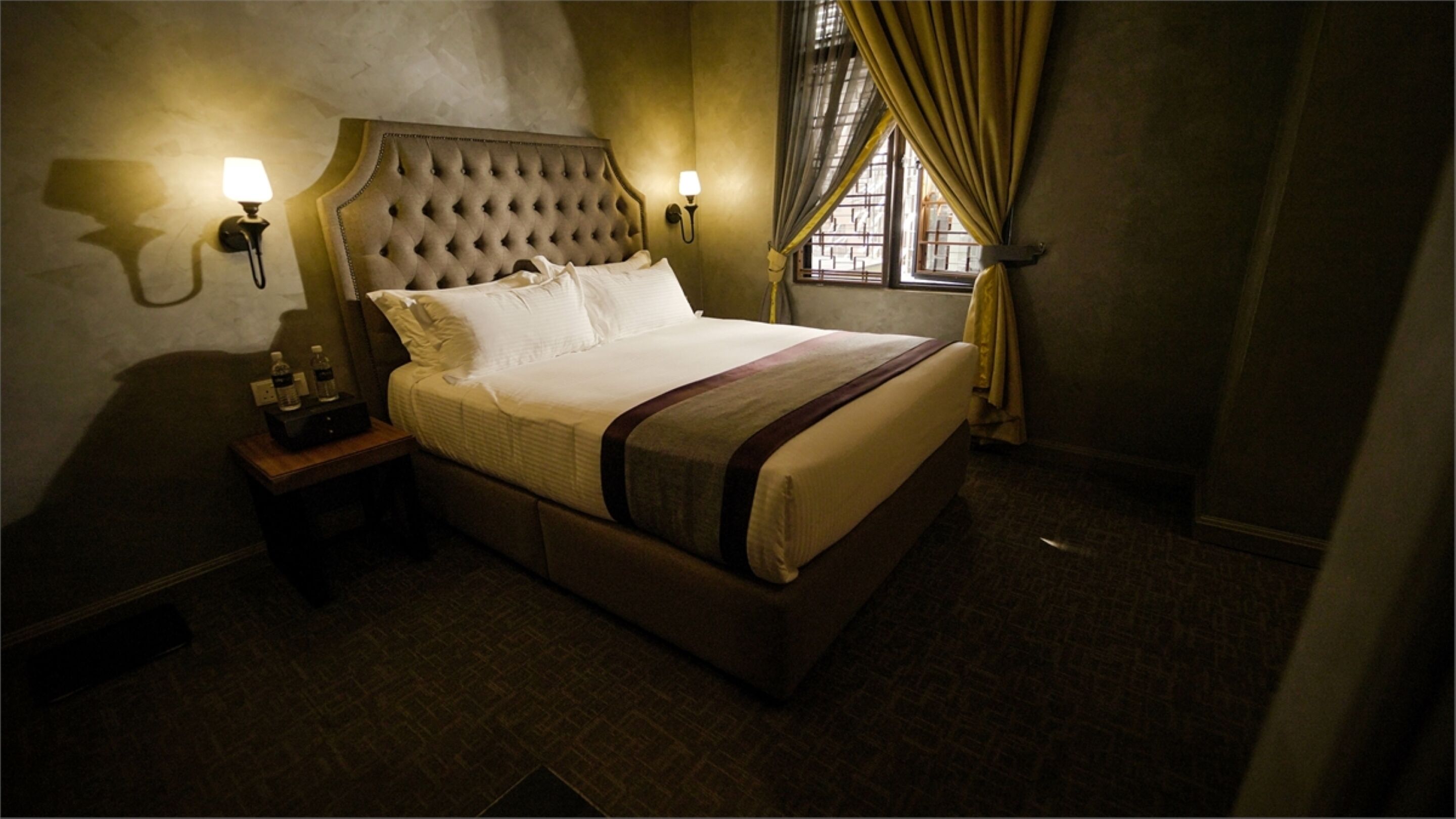 Una habitación de hotel con cabecera capitonada, una cama con ropa blanca, dos lámparas de pared, una mesita de noche con lámpara y una ventana con cortinas.
