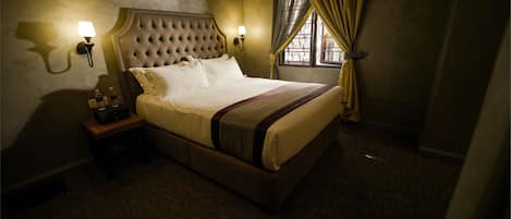 A hotel room with a tufted headboard, a bed with white linens, two wall-mounted lamps, a nightstand with a lamp, and a window with curtains.