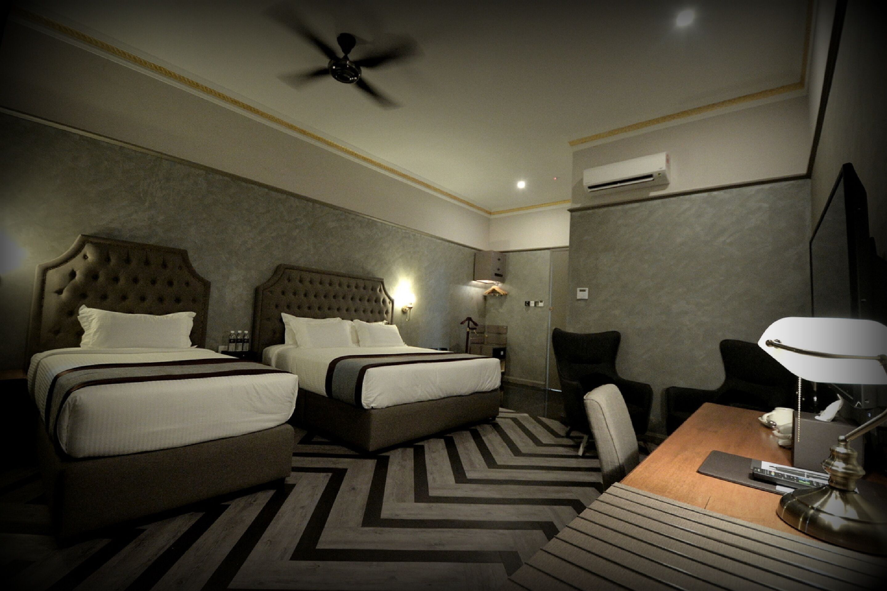 Ein Hotelzimmer mit zwei Betten, einem Schreibtisch mit Lampe, einem Sessel und einem Deckenventilator.
