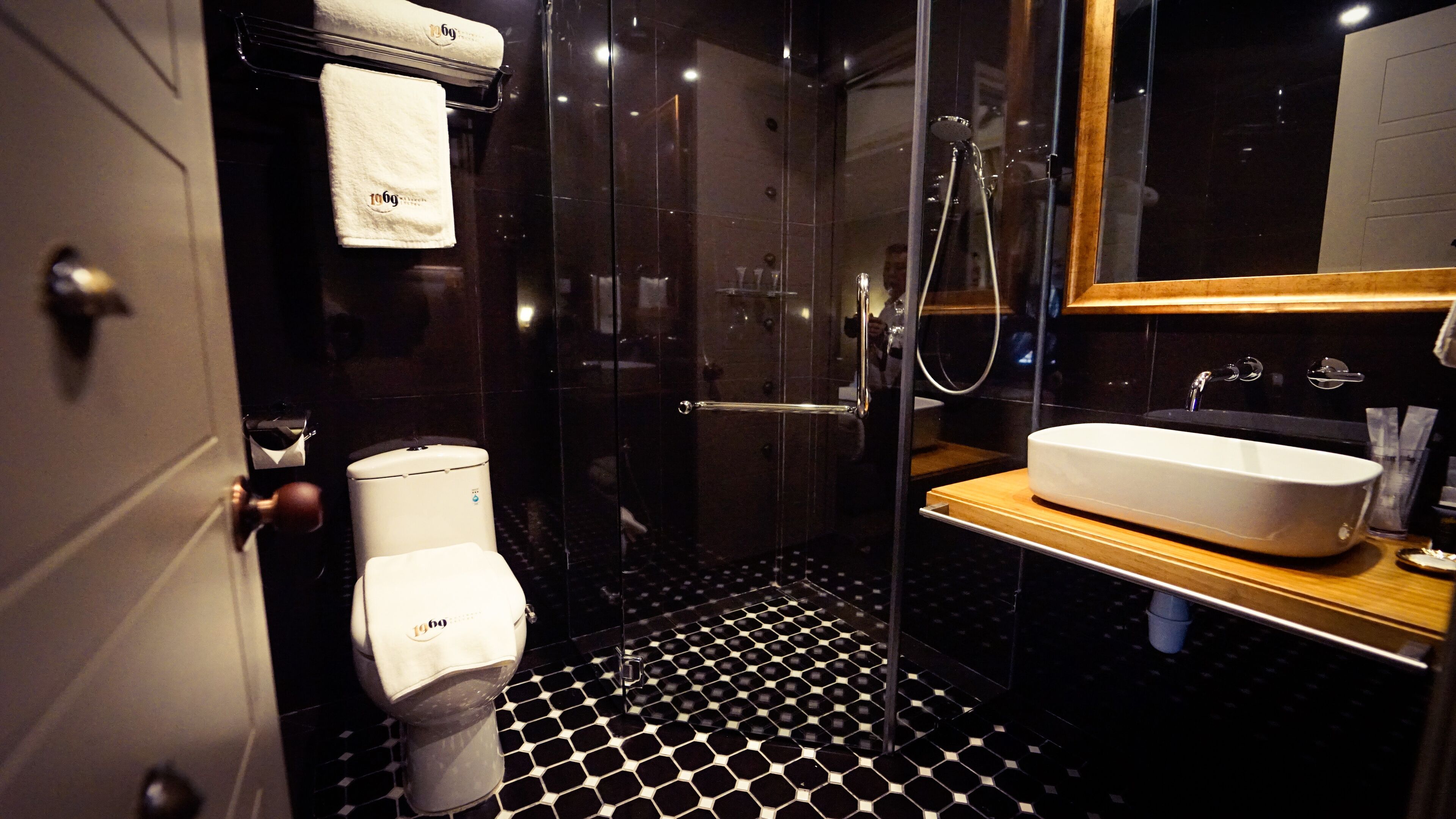 A modern bathroom with a black and white hexagonal tile floor, a white toilet, a glass-enclosed shower, and a wooden vanity with a white sink.