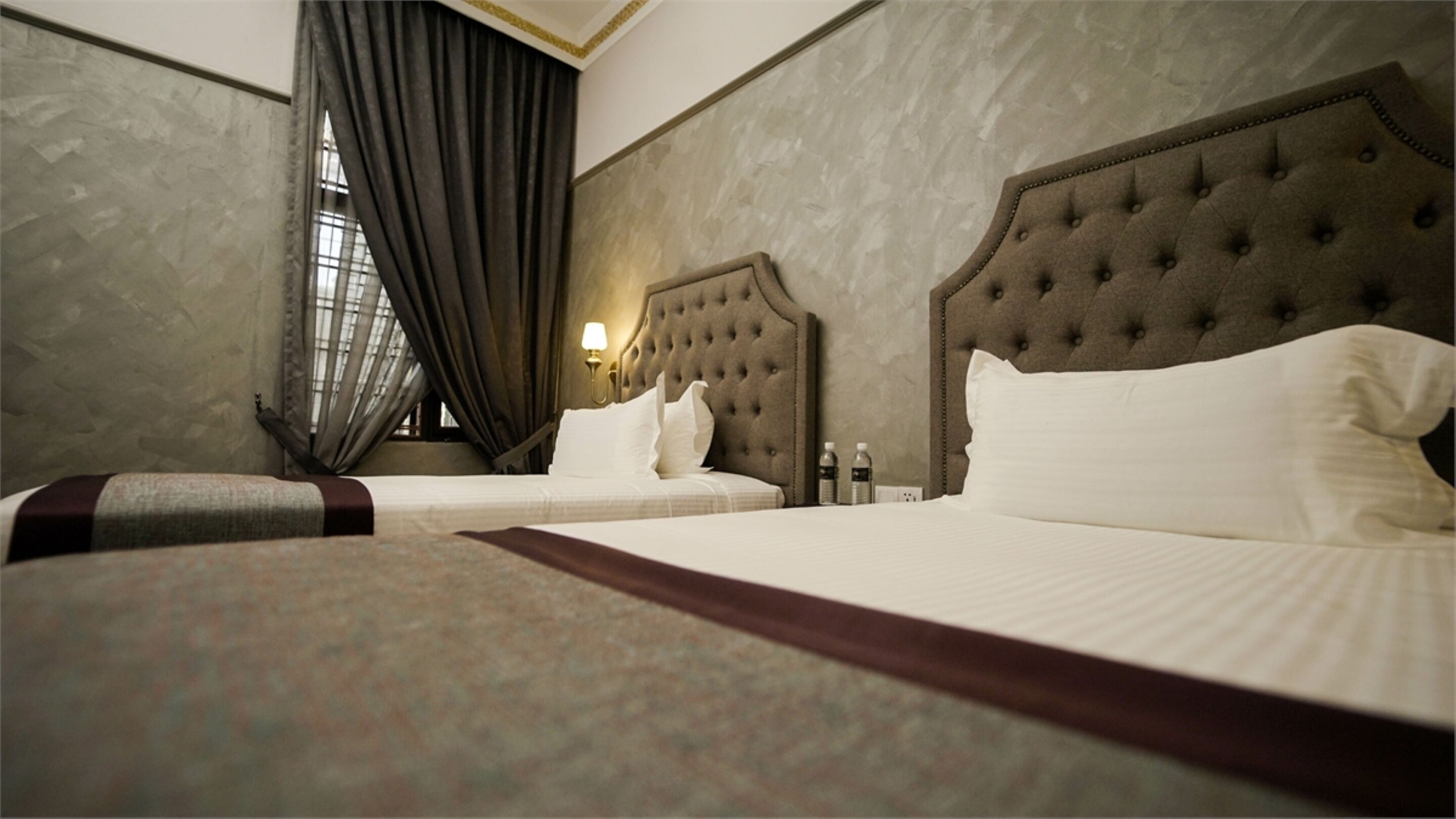 Habitación de hotel con dos camas, un ventanal grande con cortinas, una lámpara de pared y una mesita de noche con una botella.