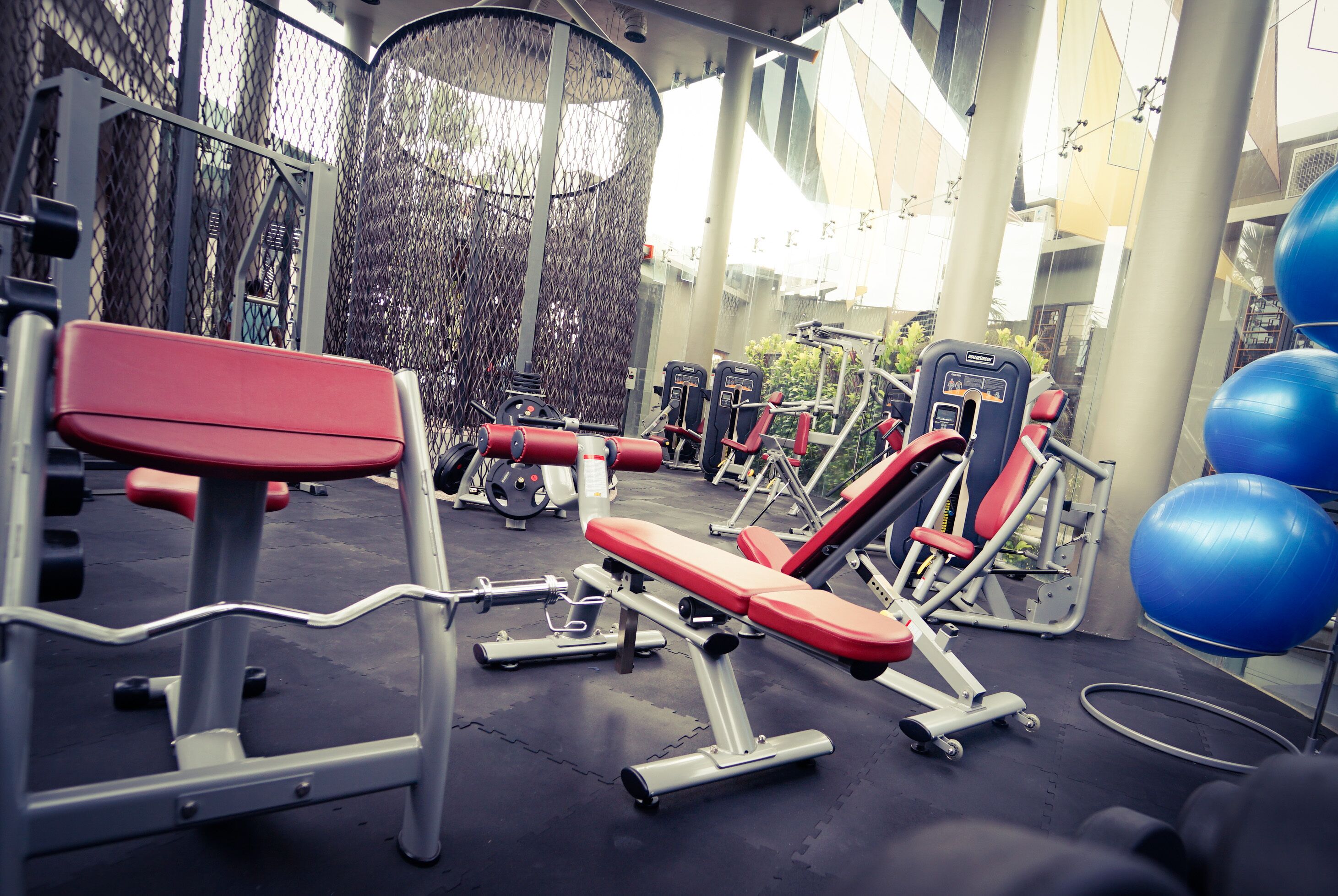 A gym with various exercise machines, including benches and weight stacks, arranged in an open space with a high ceiling and large windows.
