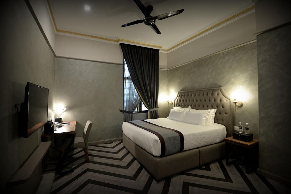 Ein Hotelzimmer mit Bett, Schreibtisch, Stuhl, Fernseher, Deckenventilator und einem Fenster mit Vorhängen.