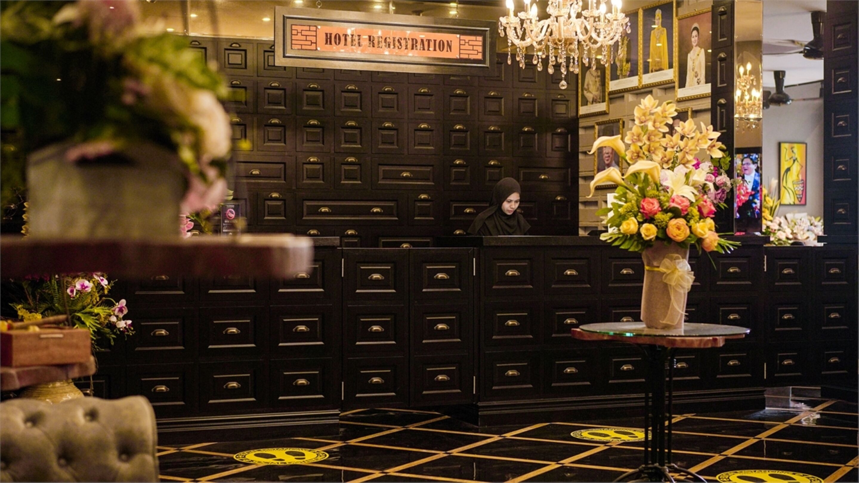 A hotel reception area with a registration desk, a chandelier, and a floral arrangement on a small table.