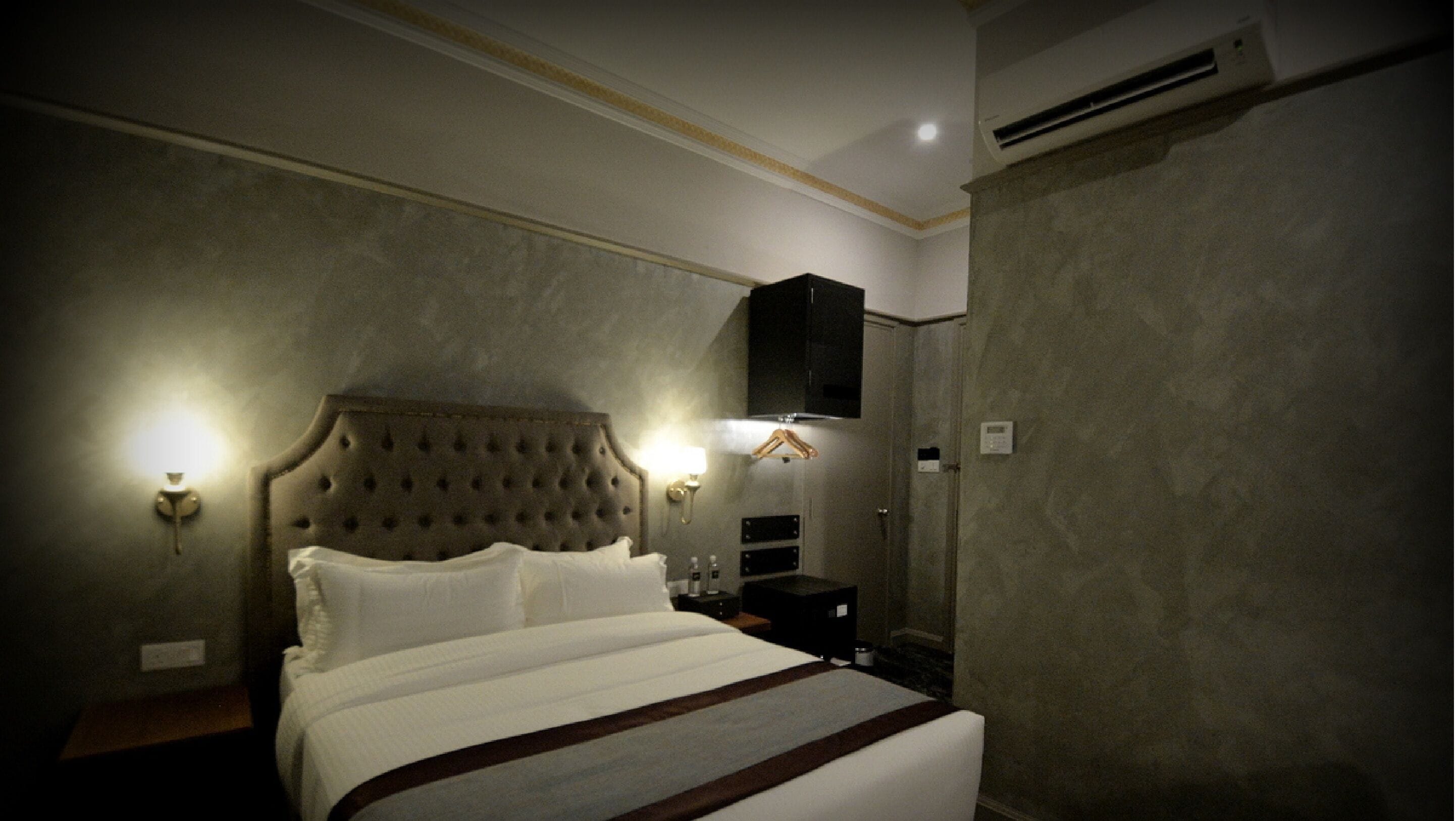 A hotel room with a tufted headboard, a large bed, bedside lamps, a wardrobe, and an air conditioning unit.