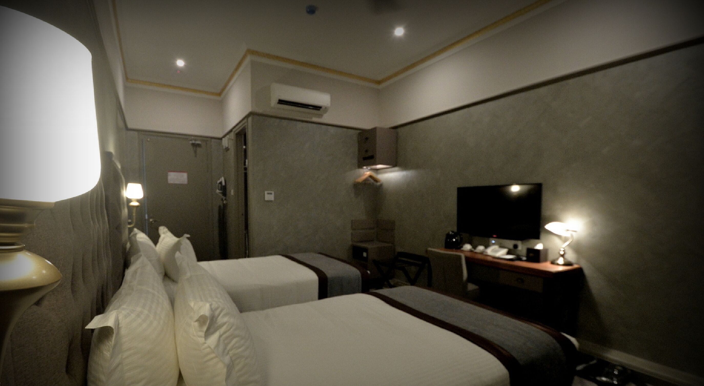 Habitación de hotel con dos camas, un escritorio con televisor, una lámpara y un aire acondicionado.
