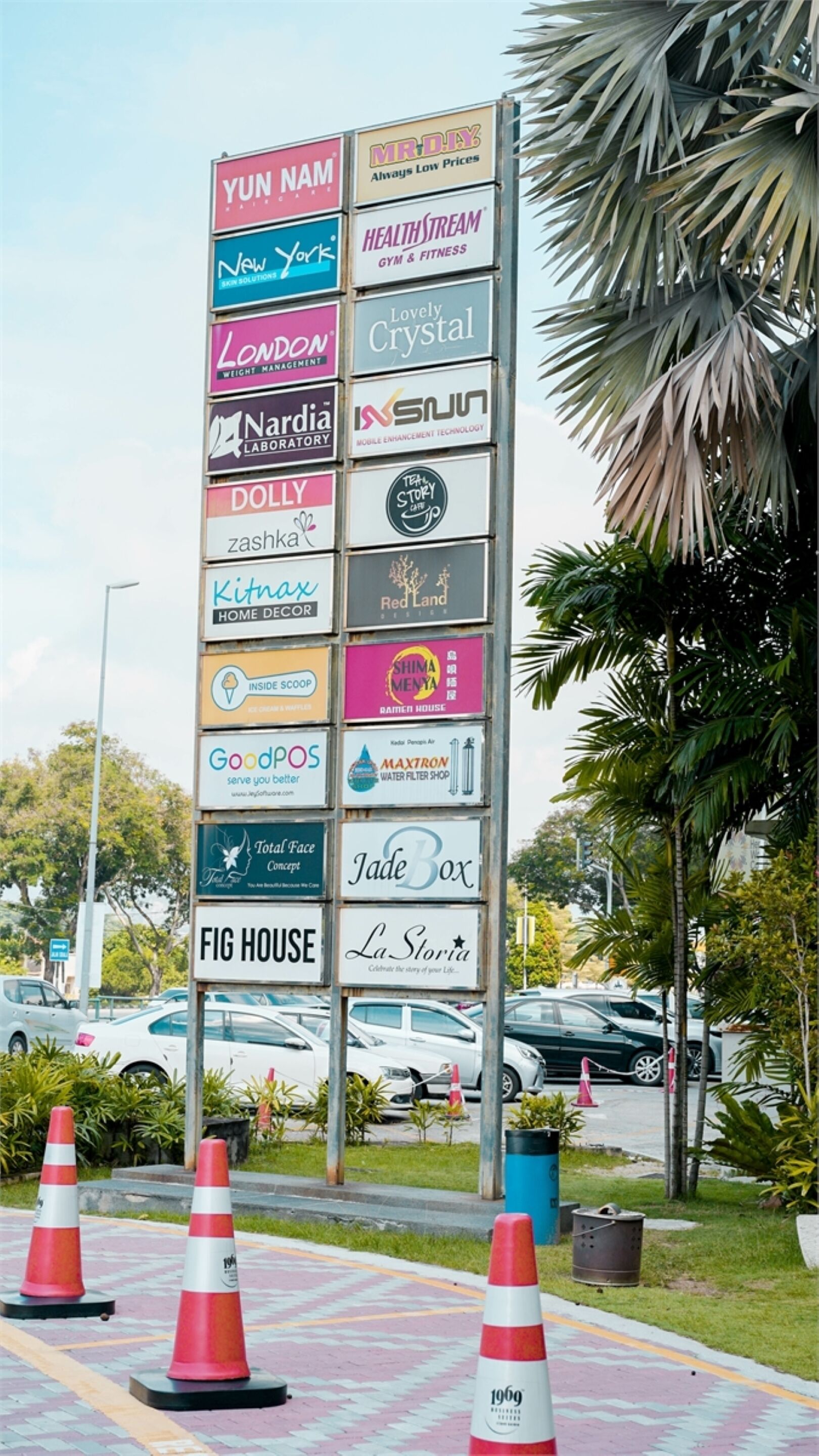 Un cartel vertical con varios logotipos de empresas, rodeado de conos de tráfico y palmeras.