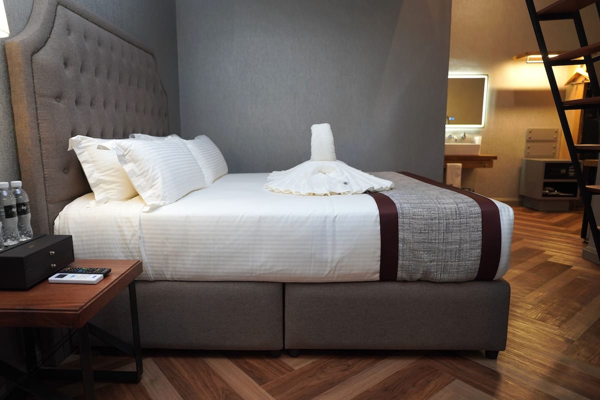 Ein ordentlich bezogenes Bett mit weißen Leinen, einem grauen Kopfteil und einer grauen Decke mit weißem Muster. Ein Holznachttisch zeigt eine Fernbedienung, eine schwarze Box und eine Wasserflasche.