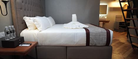 A neatly made bed with white linens, a grey headboard, and a grey and white patterned throw blanket. There is a wooden nightstand with a remote control and a black box, and a bottle of water on it.