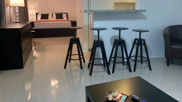A hotel room with a bed, a desk, a lamp, a TV, a ceiling fan, and a kitchenette with stools. - Miami Airport Apartment Inn (Miami)