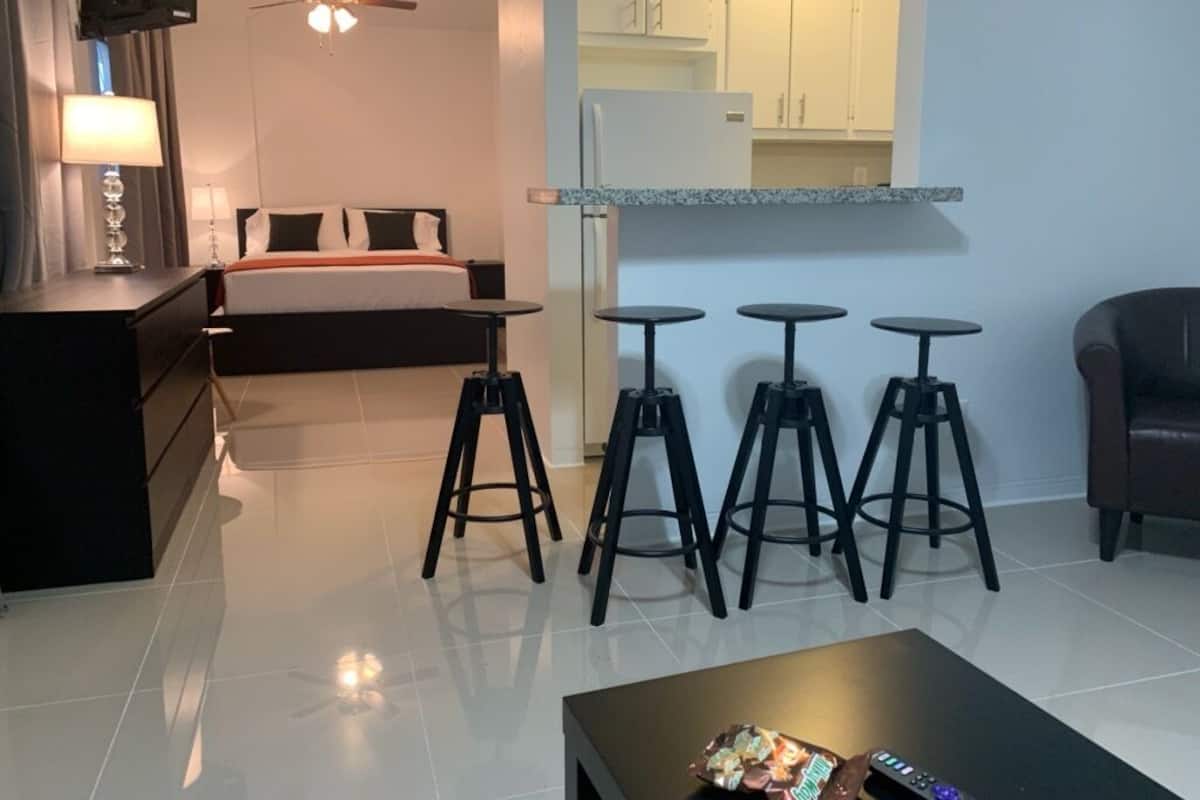 A hotel room with a bed, a desk, a lamp, a TV, a ceiling fan, and a kitchenette with stools.