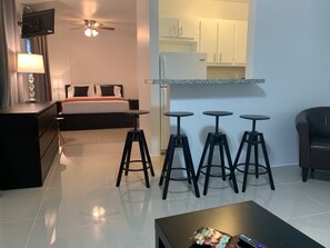 A hotel room with a bed, a desk, a lamp, a TV, a ceiling fan, and a kitchenette with stools. - Miami Airport Apartment Inn (Miami)