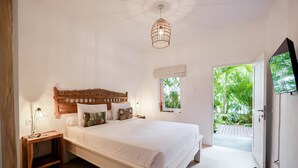 A bedroom with a wooden headboard, white bedding, a bedside lamp, and a view of greenery through an open door.