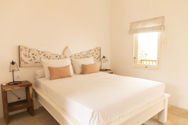A bedroom with a large bed, two bedside tables, a nightstand, a window with a blind, and a wall-mounted light fixture. - The Room Padang-Padang (Pecatu)