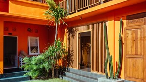 A multi-story building with a bright orange facade, a wooden balcony, and a small garden with cacti and palm plants.