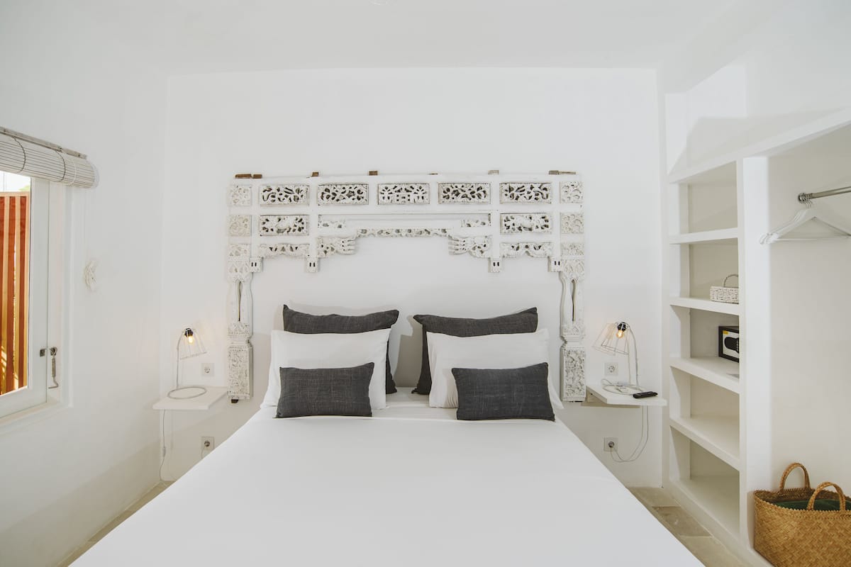 A bedroom with a white bed, two bedside tables with lamps, a decorative headboard, and a wardrobe with shelves.