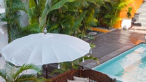 A wooden deck with a bench and a white umbrella, surrounded by lush tropical plants and a swimming pool.