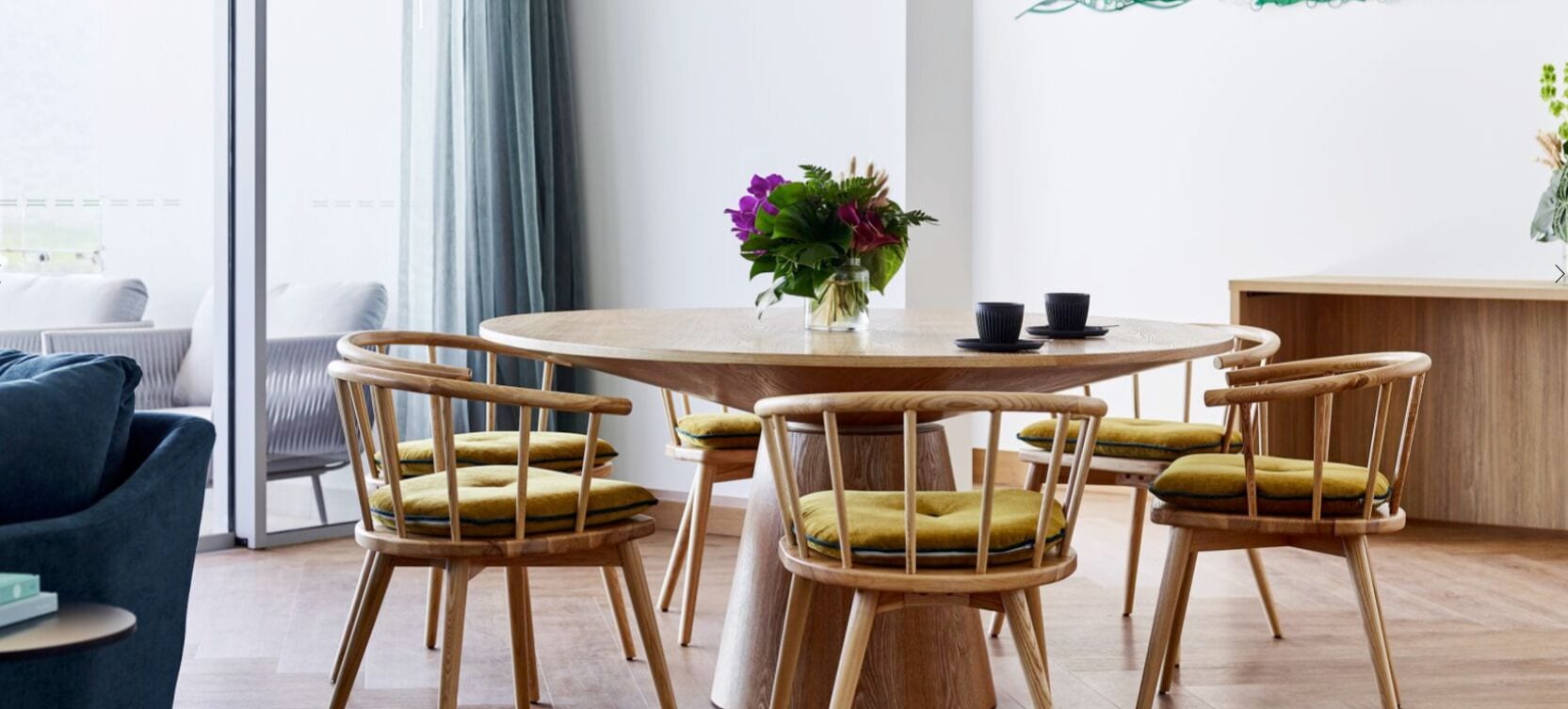 Uma mesa redonda de madeira com quatro cadeiras, cada uma com almofada. Um vaso com flores está sobre a mesa.