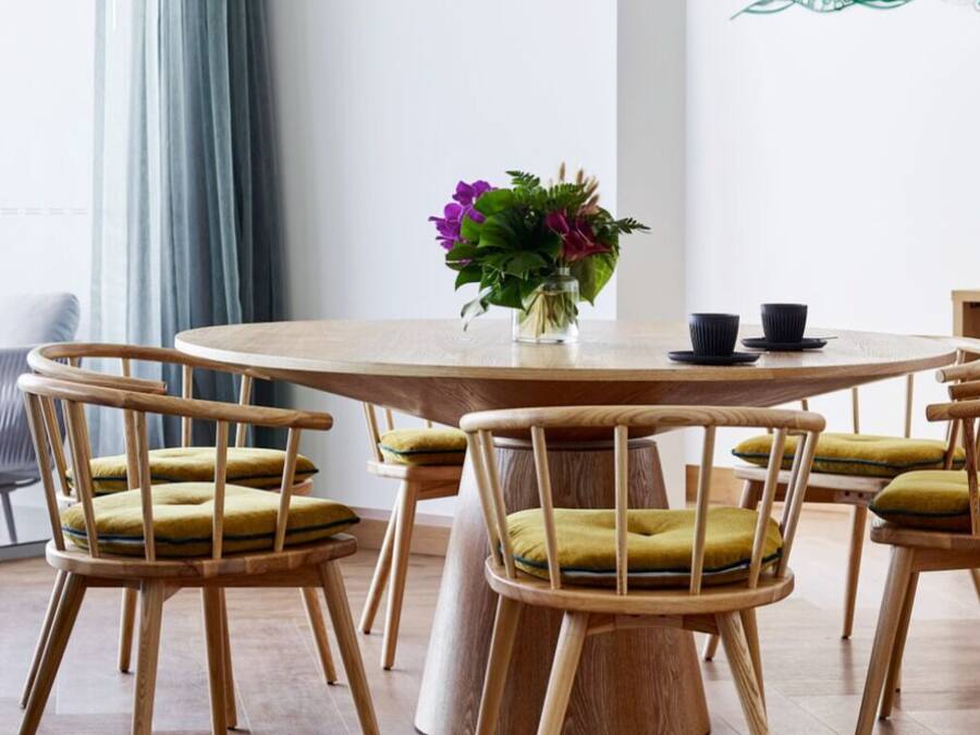 Uma mesa redonda de madeira com quatro cadeiras, cada uma com almofada. Um vaso com flores está sobre a mesa.