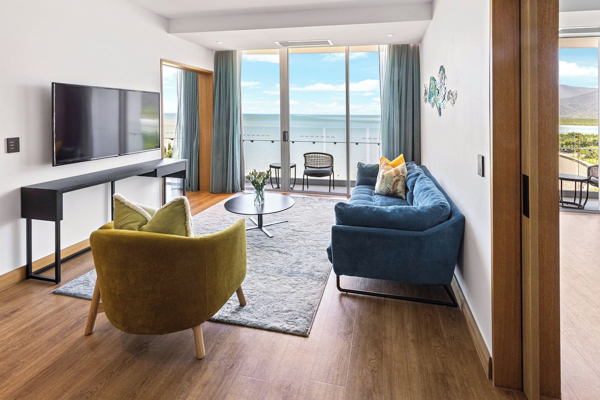 A modern hotel room with a bed, a sofa, a TV, a dining area, and a balcony with a view of the sea.