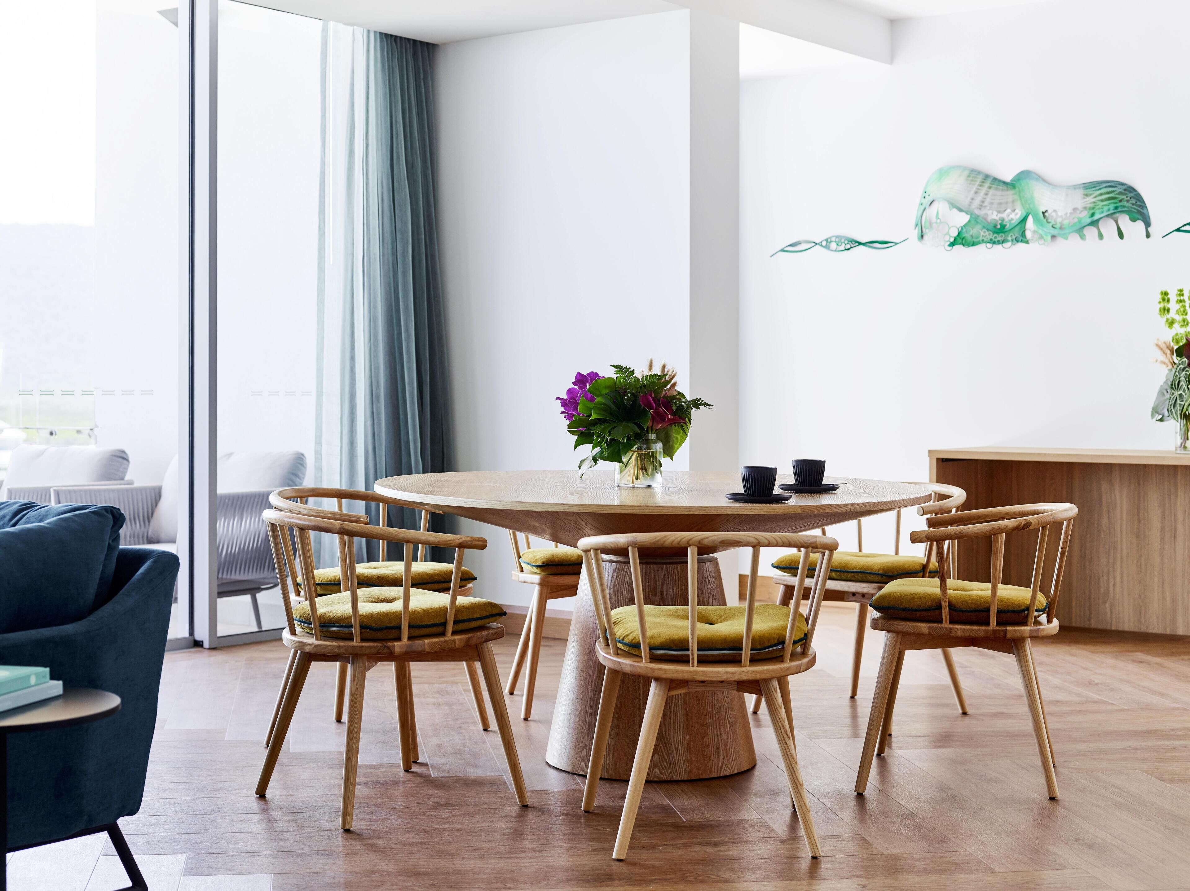 A modern dining area with a round wooden table, six wooden chairs with yellow cushions, and a vase with purple flowers.