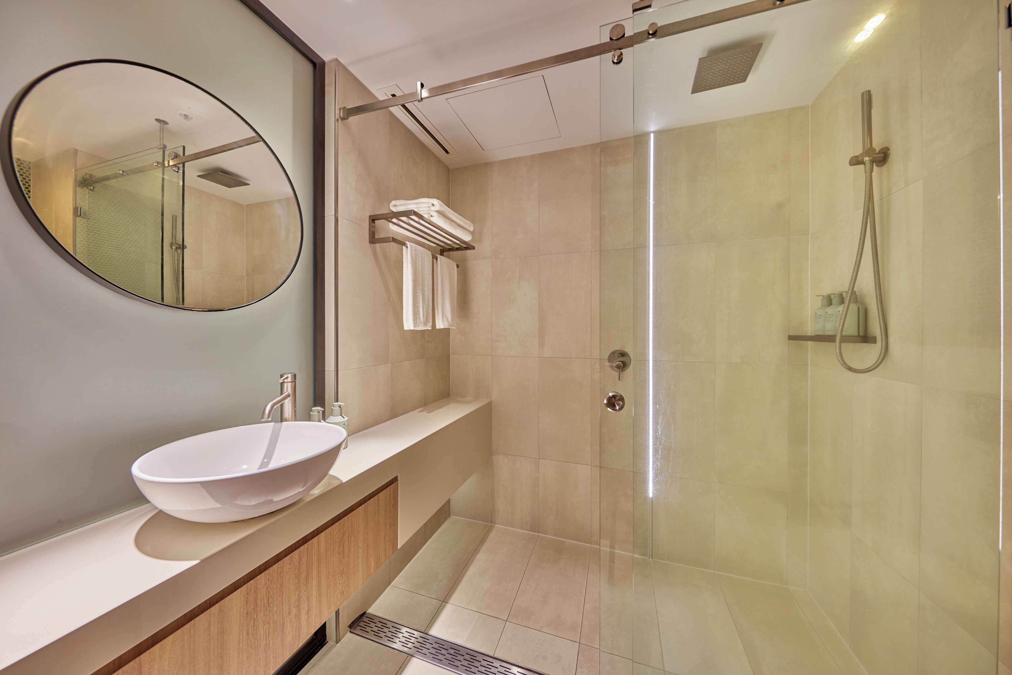 A modern bathroom with a large oval mirror, a white vessel sink, a glass-enclosed shower, and a towel rack.