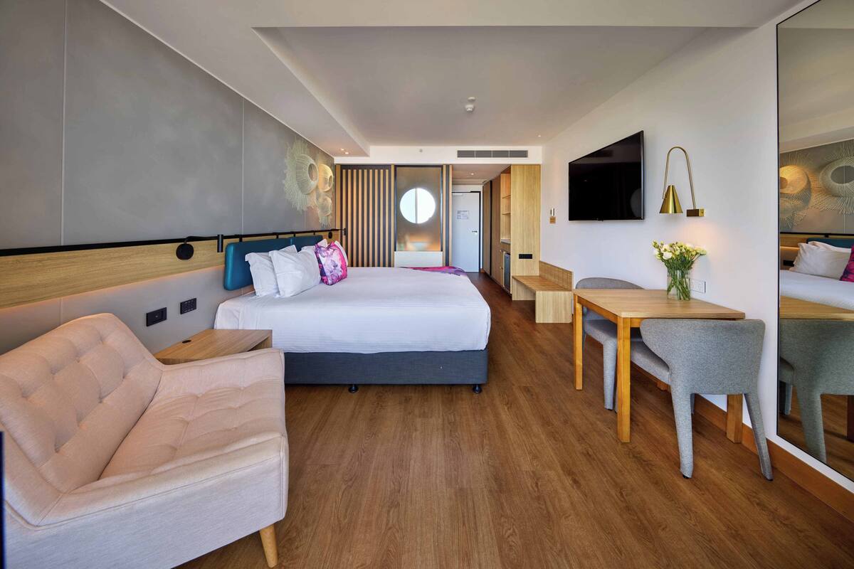 A modern hotel room with a large bed, a desk, a chair, and a TV mounted on the wall.
