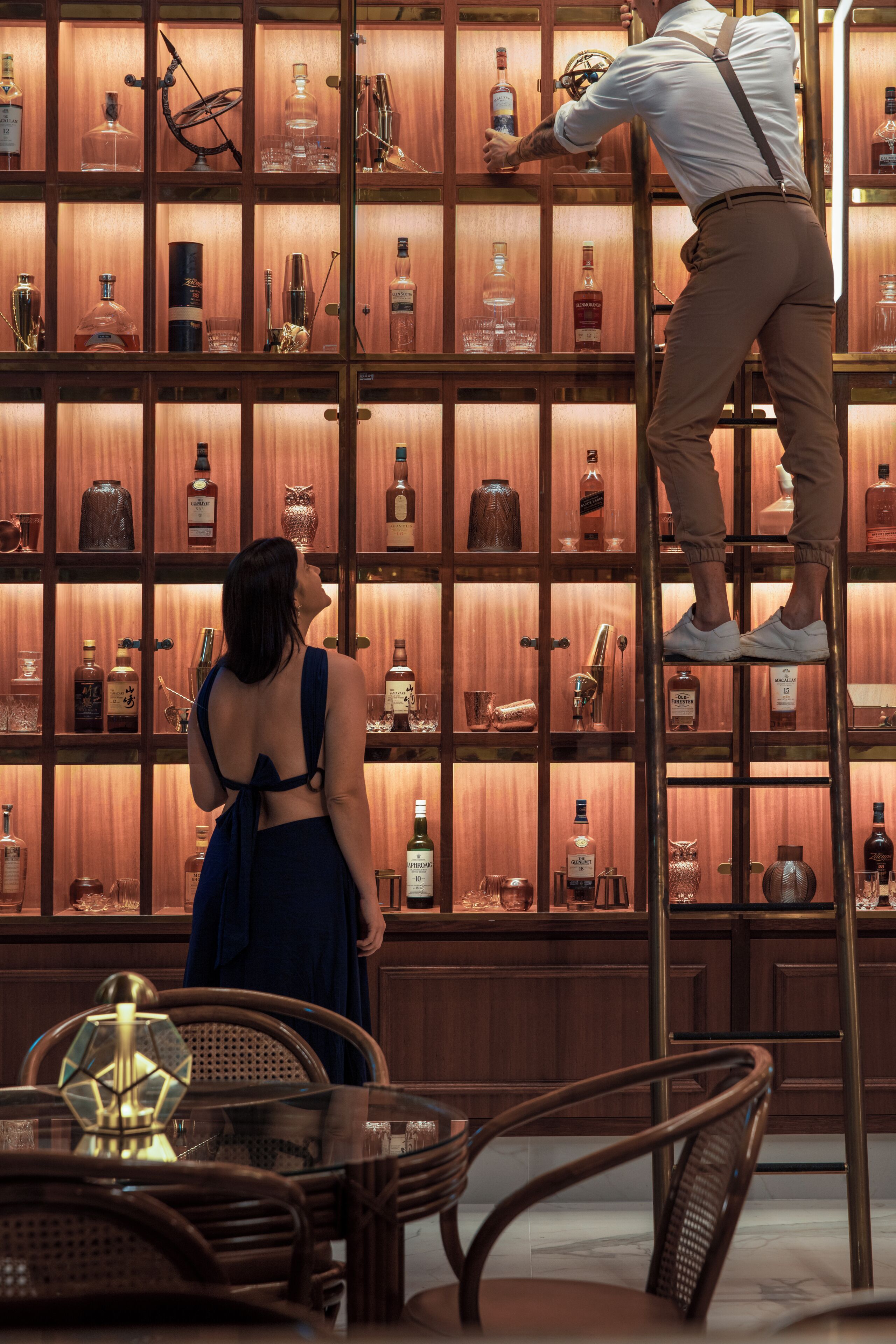 A bar area with a bartender on a ladder, a woman in a blue dress, and various bottles of liquor on shelves.