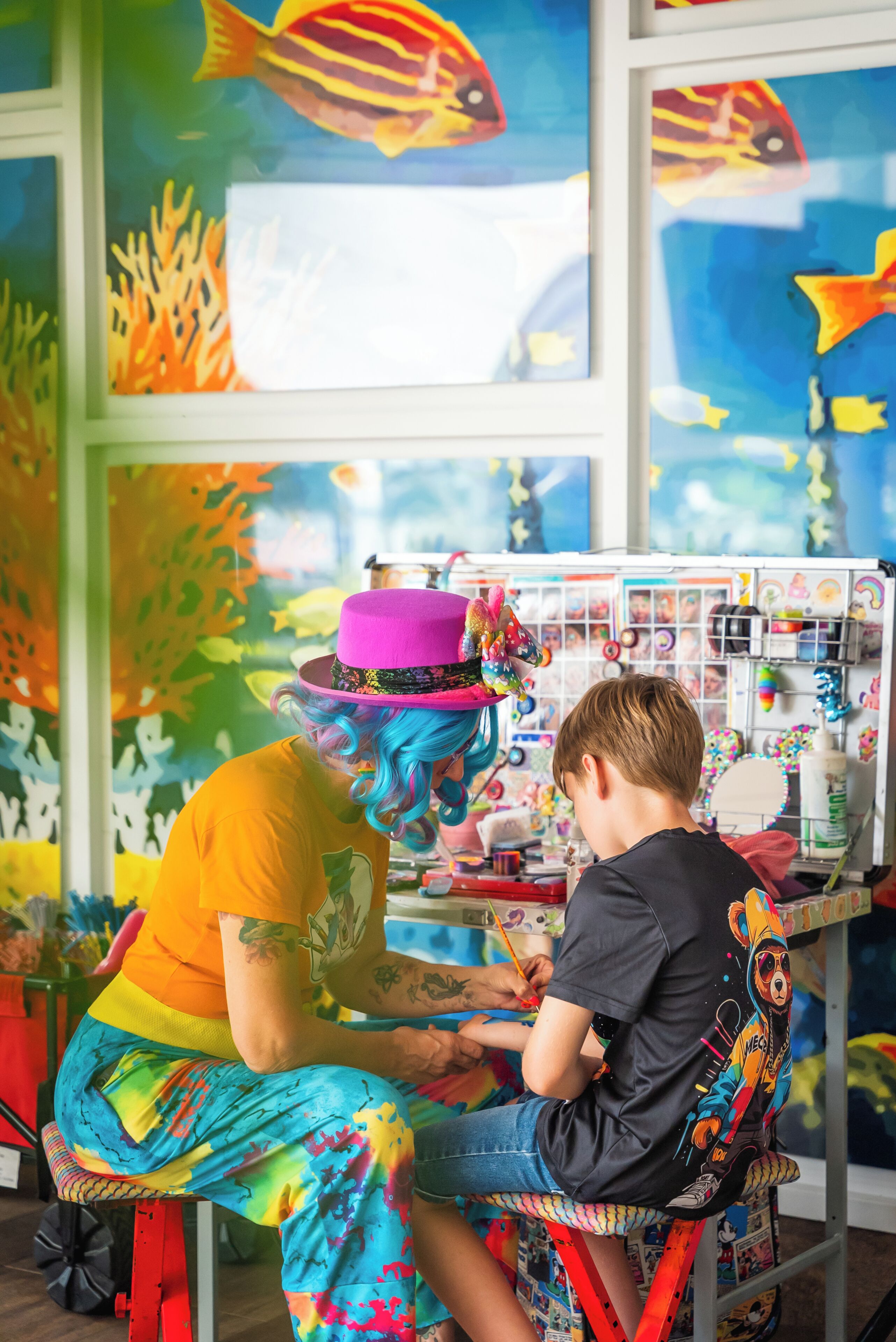 A tattoo artist working on a client's tattoo in a room with colorful murals and a window.