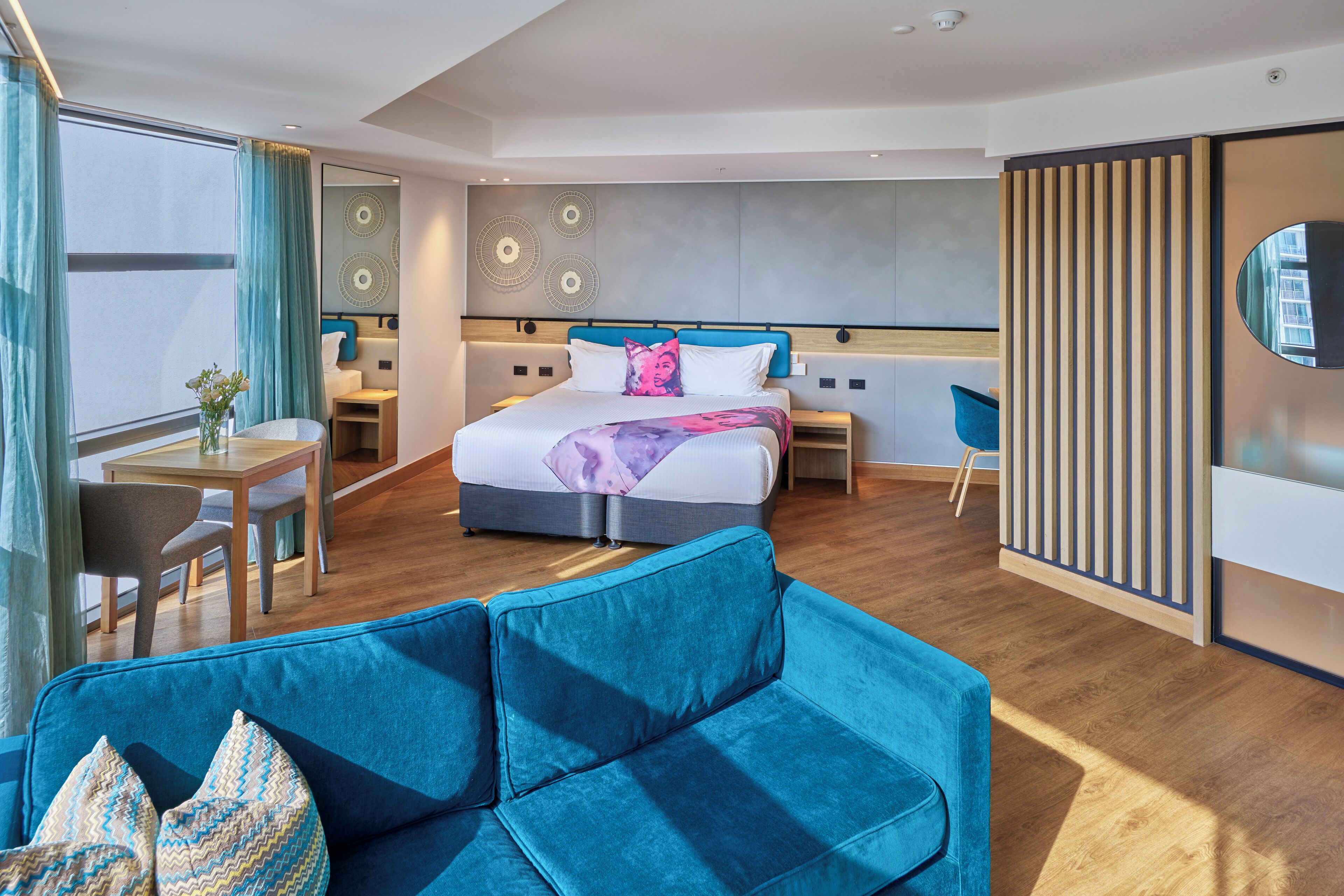 A modern hotel room with a large bed, a blue sofa, a desk, and a dining area.