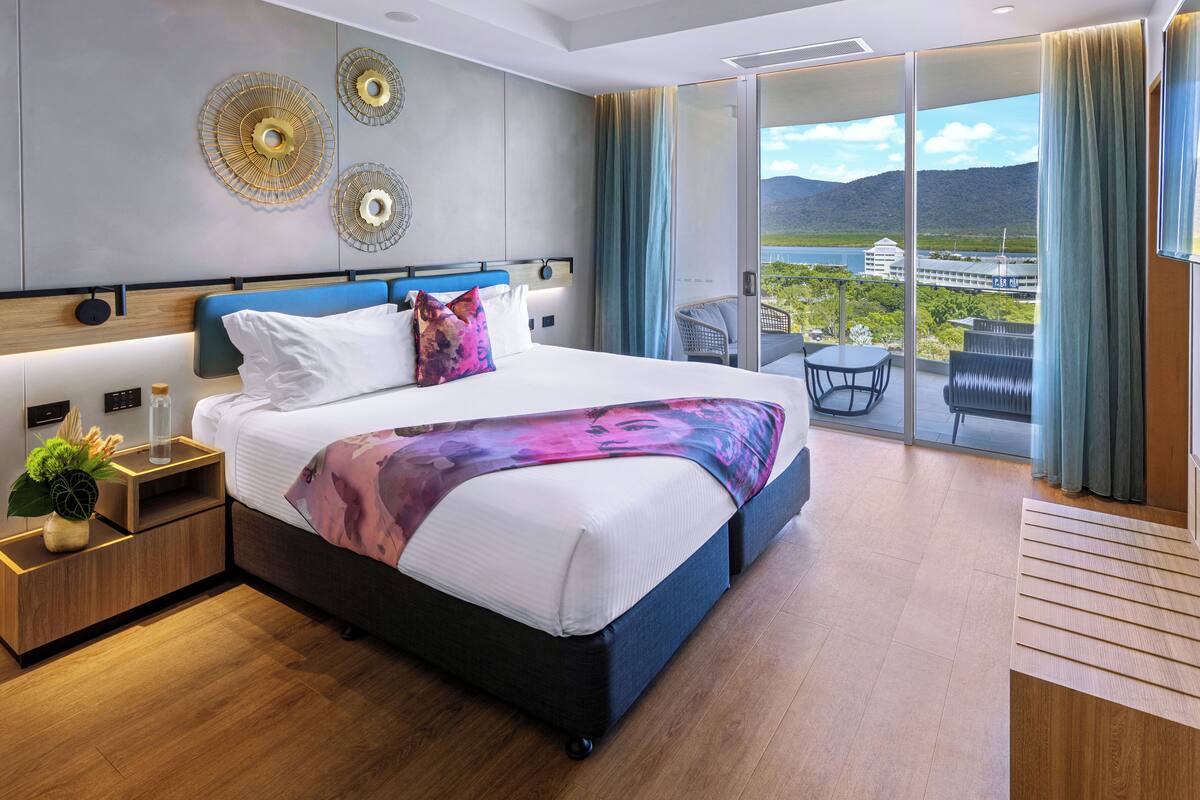 A modern hotel room with a large bed, a nightstand, a bedside table with a lamp, and a view of a balcony with a table and chairs.