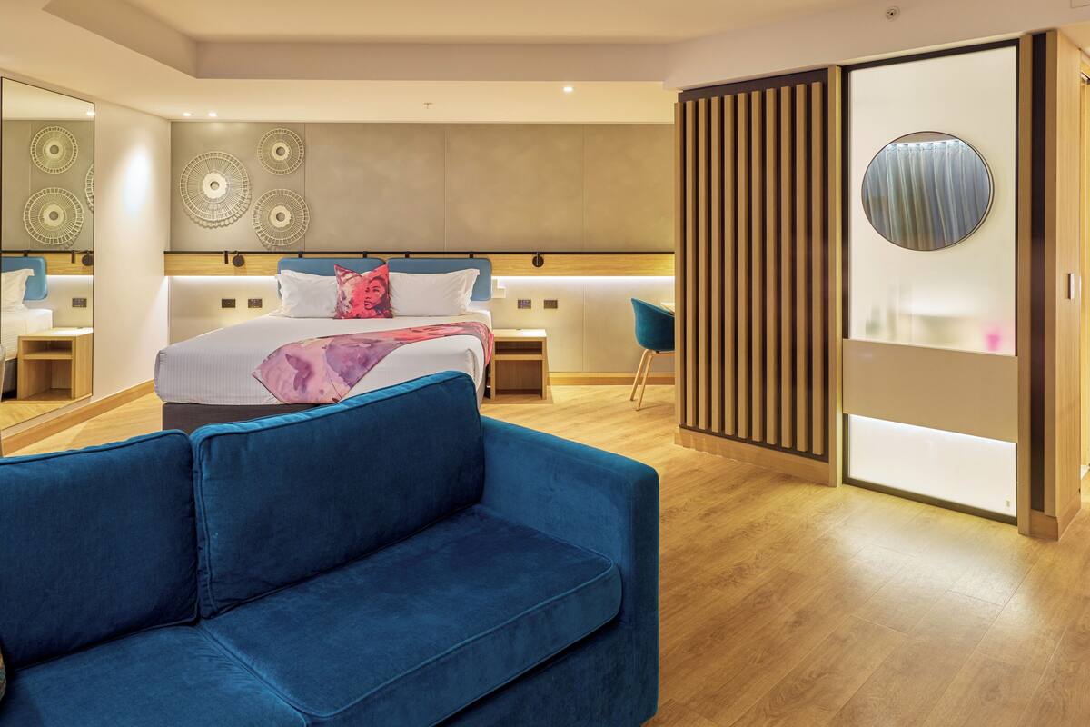 A modern hotel room with a large bed, a blue sofa, a desk, and a mirror.